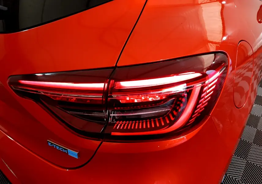 Gros plan sur le feu arrière droit allumé d'une Renault Clio Initiale Paris E-TECH orange 2021 avec logo hybride visible.