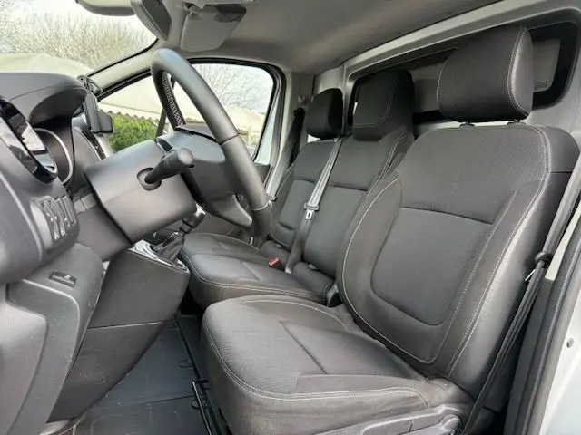 Vue intérieure côté conducteur du Renault Trafic Fourgon gris platine, montrant les sièges avant en tissu noir et le volant.