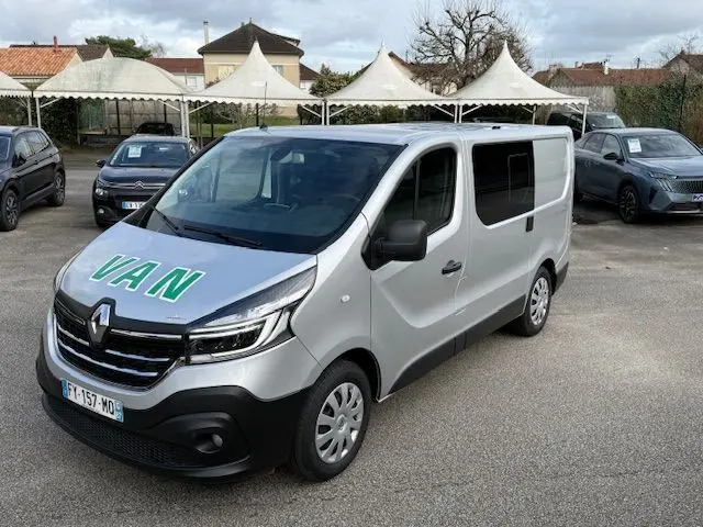 Vue 3/4 avant d’un Renault Trafic Fourgon gris platine avec inscription VAN en vert sur le capot, stationné en extérieur.
