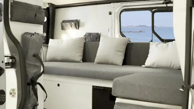 Vue intérieure du Renault Trafic Fourgon gris platine, montrant l'aménagement avec banquette grise et coussins clairs près d'une fenêtre ouverte sur la mer.