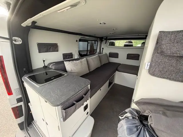 Vue intérieure arrière droite du Renault Trafic Fourgon gris platine aménagé avec banquette convertible, rangements et kitchenette compacte.
