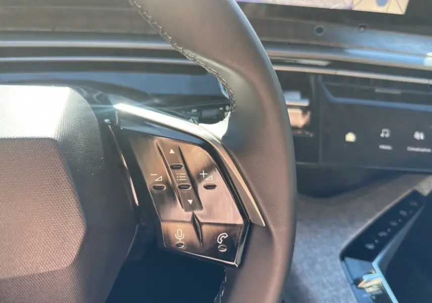 Gros plan sur les commandes du volant cuir multifonctions du Peugeot 5008 Hybrid 145 gris Artense, tableau de bord numérique en arrière-plan.