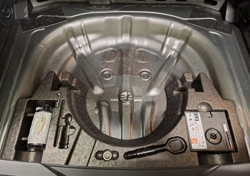 Coffre ouvert d'un Volkswagen T-Roc 2024 gris Indium montrant le rangement pour roue de secours et outils intégrés.