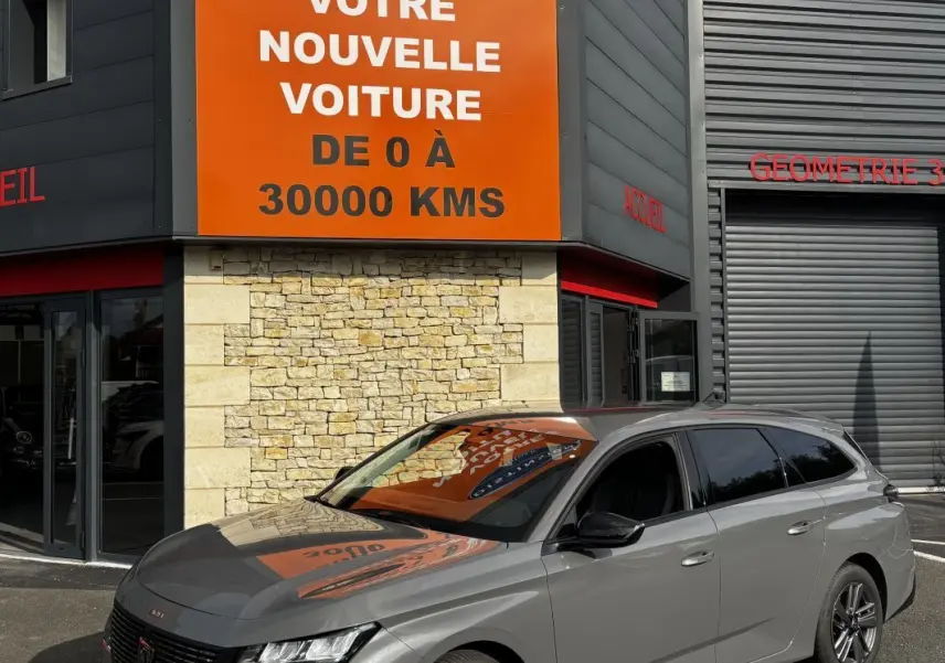 Peugeot 308 PureTech 130 S&S Allure gris clair vue 3/4 avant droit devant un bâtiment avec panneau orange.