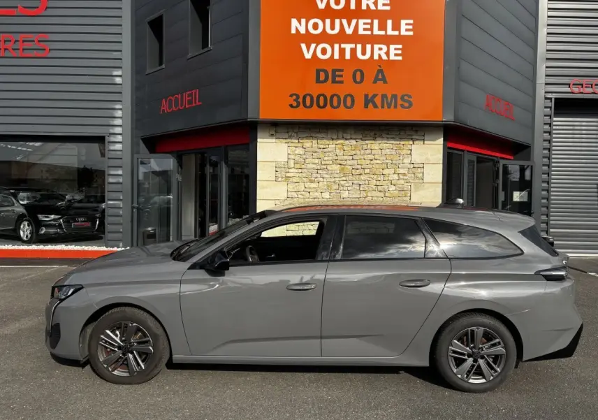 Profil côté gauche d'une Peugeot 308 PureTech 130 S&S Allure gris clair 2024 stationnée devant un bâtiment commercial.