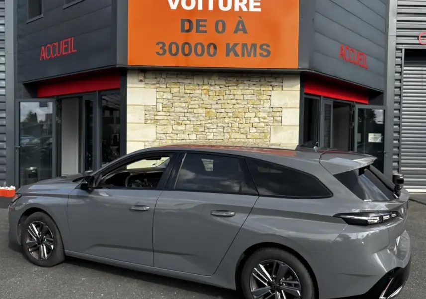 Peugeot 308 PureTech 130 gris clair vue de profil côté gauche, break garé devant un bâtiment avec panneau orange.