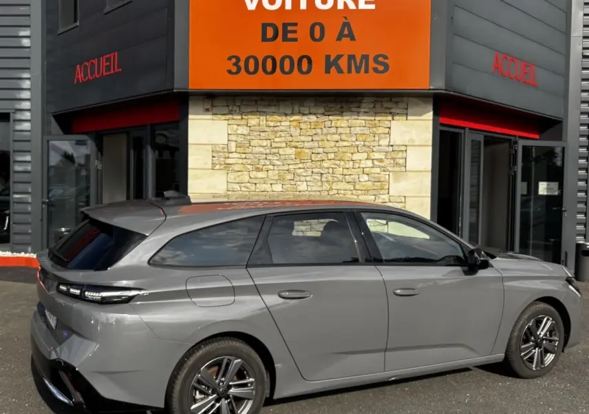 Peugeot 308 PureTech 130 S&S Allure gris clair vue de profil côté gauche devant un bâtiment avec panneau orange "Votre nouvelle voiture".