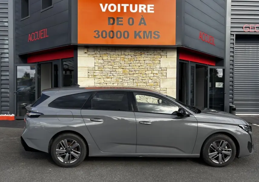 Peugeot 308 PureTech 130 S&S Allure gris clair vue de profil côté gauche devant un bâtiment avec panneau orange.