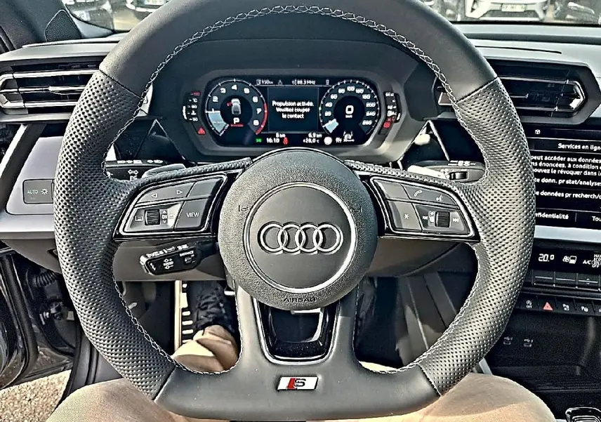 Vue rapprochée du volant sport en cuir noir avec logo Audi et badge S, tableau de bord digital en arrière-plan.
