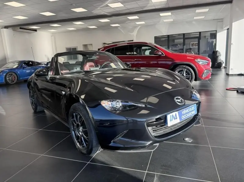 Mazda MX-5 noir 2020 en 3/4 avant droit, capote ouverte, intérieur rouge visible dans un showroom lumineux.