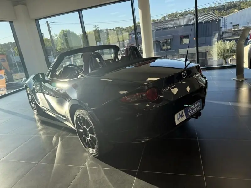 Mazda MX-5 noir 2020 vue 3/4 arrière côté gauche, capote ouverte, dans un showroom lumineux.