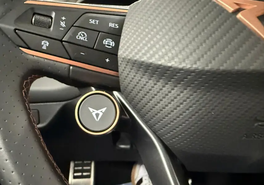 Gros plan sur le volant en carbone et cuir perforé avec logo CUPRA et commandes d'assistance au conducteur.