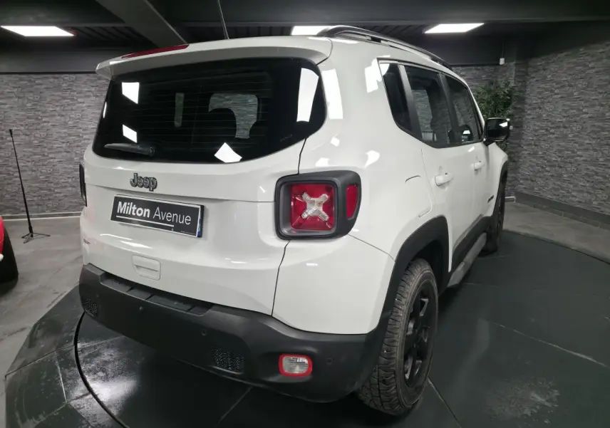 Vue 3/4 arrière droite d'un Jeep Renegade blanc avec jantes noires 5 branches et barres de toit gris clair.