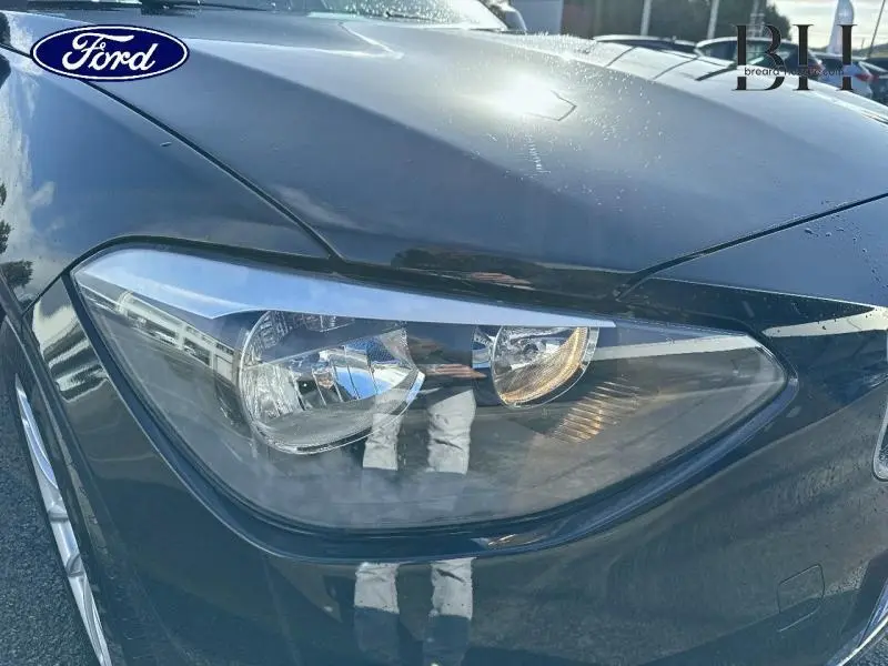 Gros plan sur le phare avant gauche d'une BMW Série 1 114i noire, avec reflets sur la carrosserie brillante.