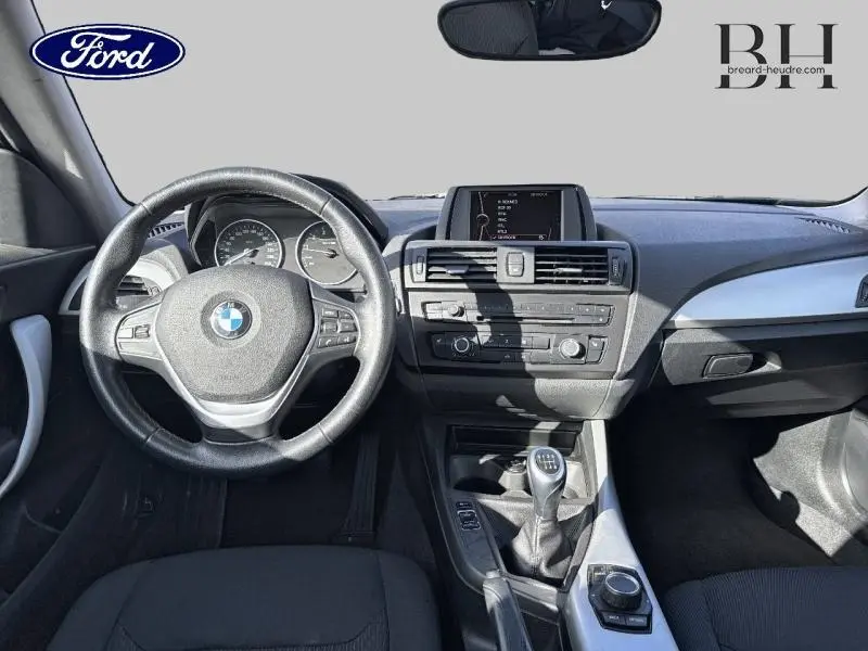 Vue intérieure avant de la BMW Série 1 114i noire de 2013, volant cuir, tableau de bord et boîte manuelle visibles.