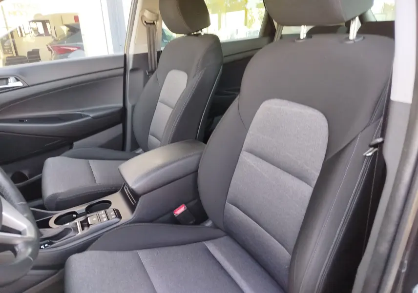 Vue intérieure côté conducteur du Hyundai Tucson 2019, sièges tissu gris foncé avec accoudoir central noir.