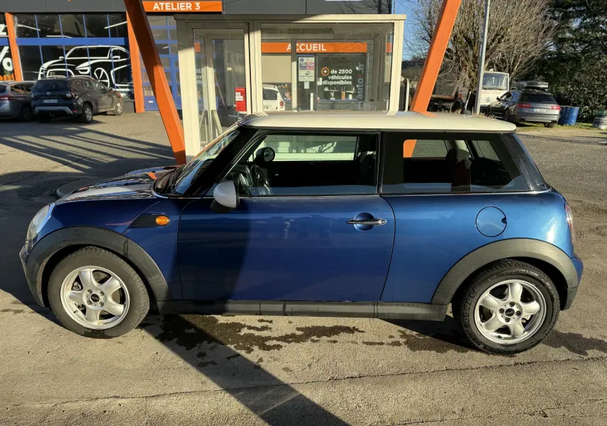 Profil côté gauche d'une MINI Cooper 1.6 bleu avec toit blanc, garée devant un atelier automobile.