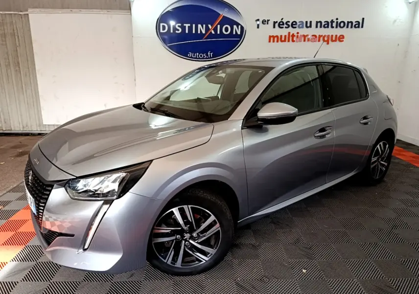 Peugeot 208 gris clair vue 3/4 avant droit, avec feux LED et jantes alliage dans un showroom intérieur.