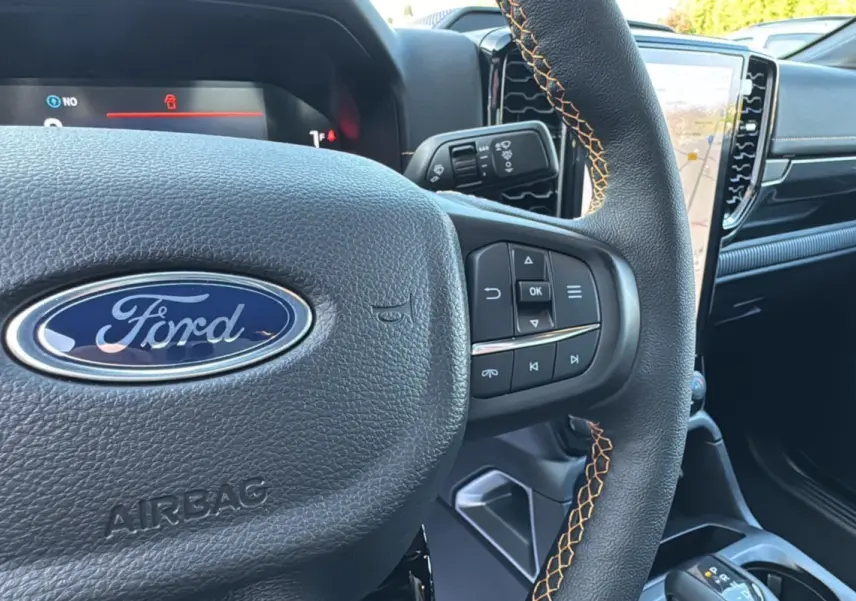 Gros plan sur le volant cuir noir avec surpiqûres orange du Ford Ranger 2.3 PHEV, tableau de bord et écran tactile visibles.