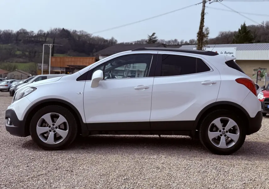 Profil côté gauche d'un Opel Mokka X blanc 2016 avec jantes alliage et toit noir, stationné sur un sol gravillonné.