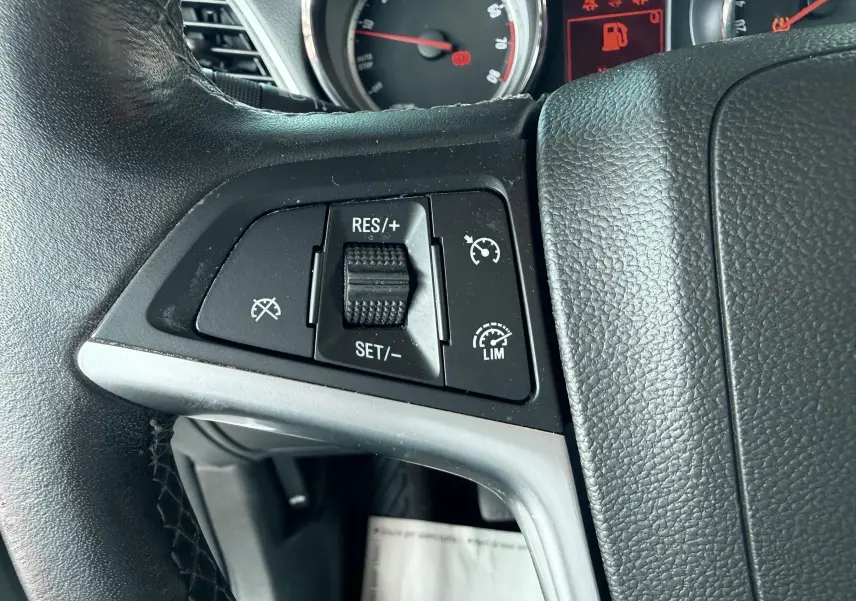 Gros plan sur les commandes de régulateur de vitesse au volant noir d’un Opel Mokka X blanc, tableau de bord en arrière-plan.