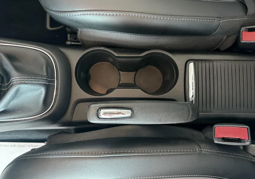 Vue plongeante sur la console centrale noire de l'Opel Mokka X avec frein à main, porte-gobelets et sièges en cuir noir.