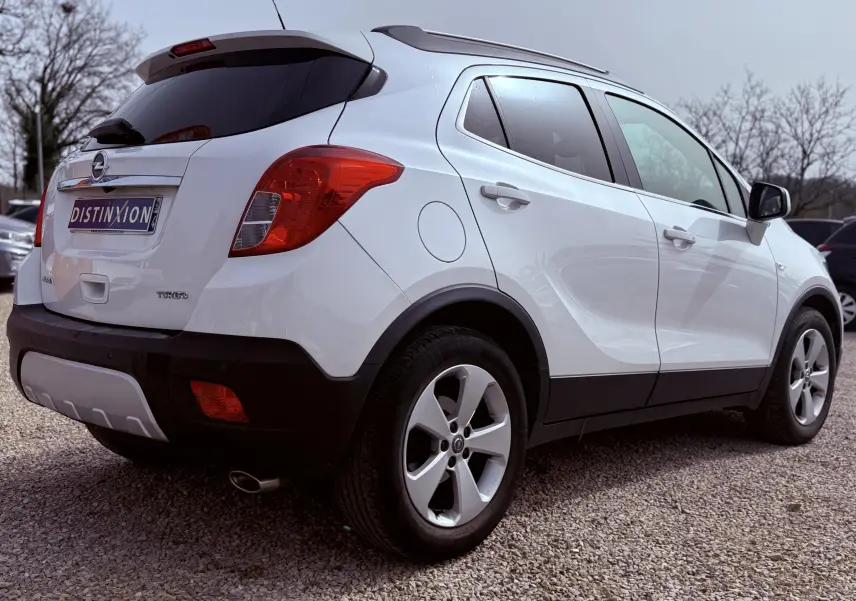 Vue 3/4 arrière droite d'un Opel Mokka X blanc avec jantes alliage et vitres teintées sur un sol gravillonné.