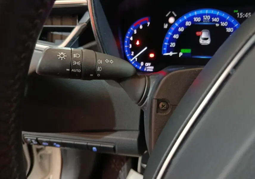 Gros plan sur le combiné d'instruments numérique bleu et le commodo d'éclairage à gauche du volant dans une Toyota Corolla hybride blanche.