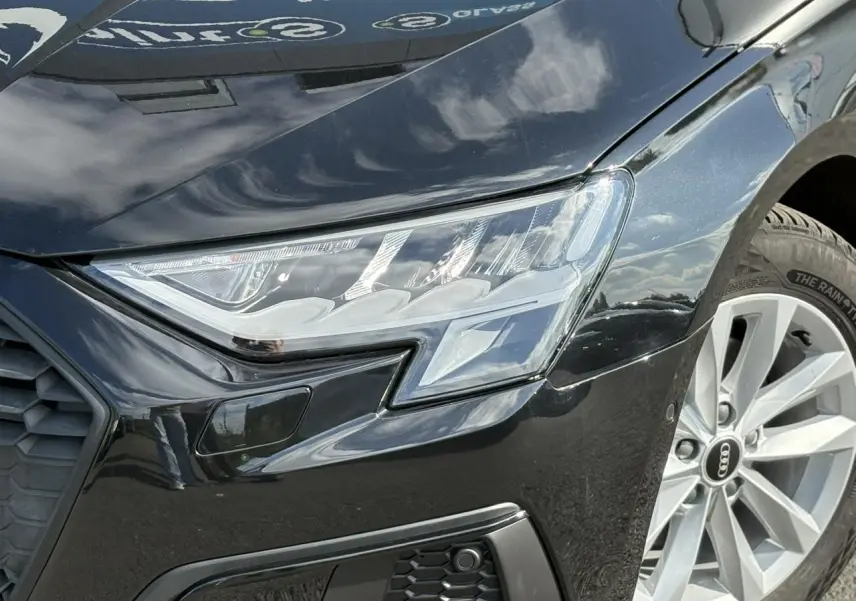 Gros plan sur le phare avant droit et la roue alliage 16 pouces d'une Audi A3 Sportback 30 TFSI noire Mythe 2023.