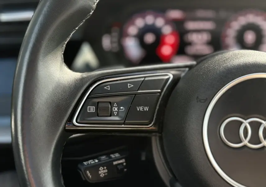 Gros plan sur le volant noir de l'Audi A3 Sportback 30 TFSI 2023 avec commandes multifonctions et affichage numérique flou.