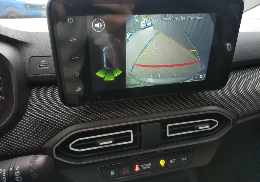 Écran central de la Dacia Sandero 2024 gris montrant la caméra de recul avec guidage de trajectoire et capteurs d’aide au stationnement.