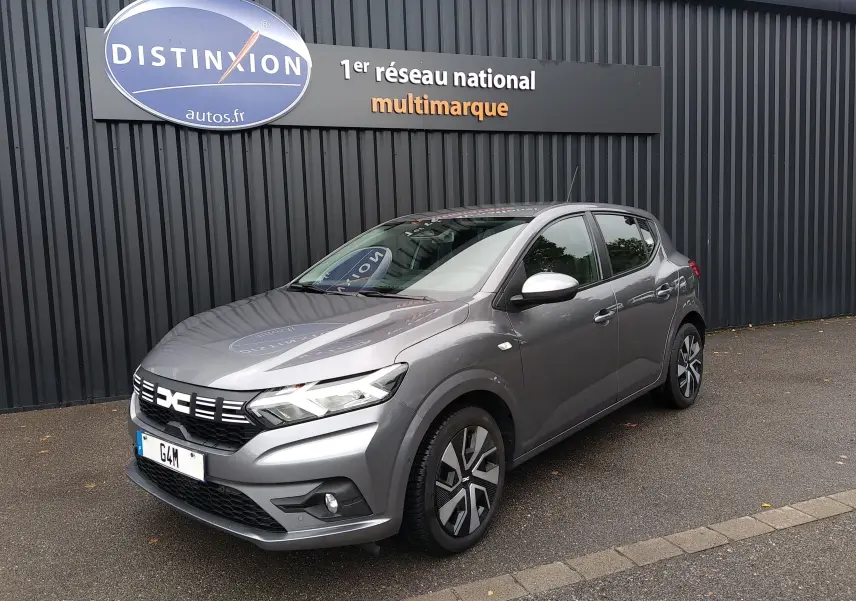 Dacia Sandero 1.0 ECO-G gris vue 3/4 avant droit, avec jantes distinctives et calandre moderne, stationnée devant un mur noir.