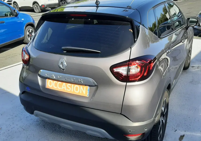 Vue 3/4 arrière droite d'un Renault Captur gris Cassiope avec toit noir, en vente sur parking automobile.