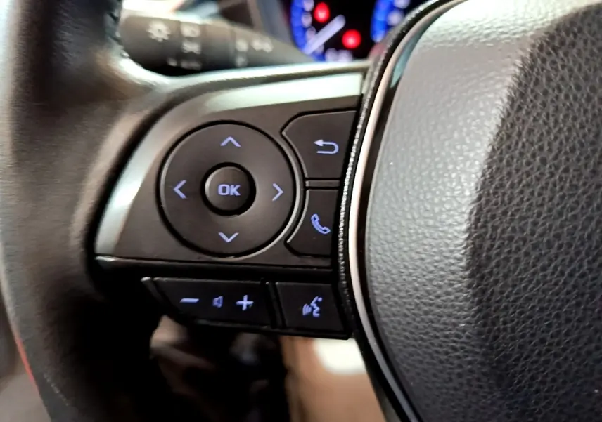 Gros plan sur le volant cuir et alu du Suzuki Swace gris clair, avec commandes multifonctions rétroéclairées en bleu.