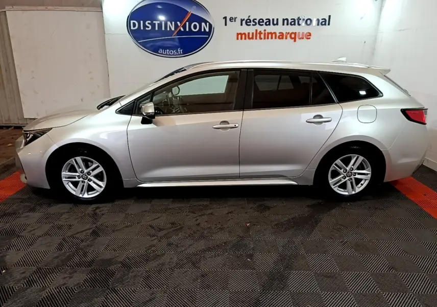 Profil droit d'une Suzuki Swace gris clair 2021 en version hybride, avec jantes alliage et toit chromé visible en intérieur showroom.