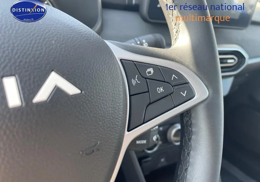 Gros plan sur les commandes au volant de la Dacia Sandero gris schiste, avec tableau de bord et aérateurs visibles en arrière-plan.