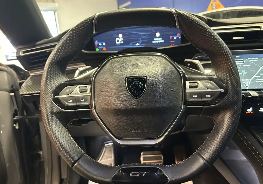 Volant cuir perforé avec logo Peugeot et badge GT, tableau de bord digital et écran tactile en arrière-plan.