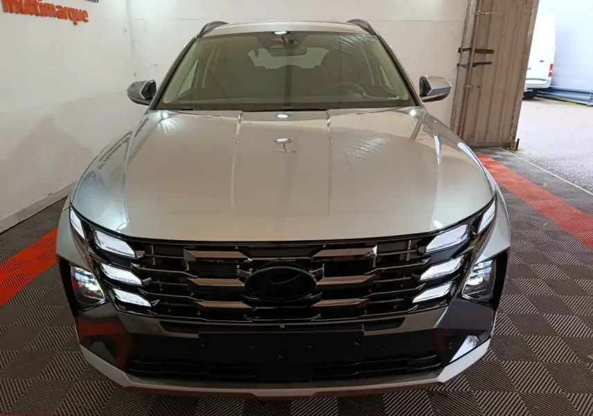 Vue avant d’un Hyundai Tucson gris clair 2025 avec calandre large et phares LED intégrés, en intérieur showroom.