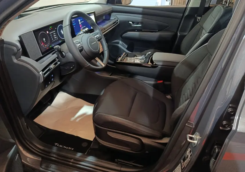 Vue intérieure côté conducteur du Hyundai Tucson gris clair 2025, avec tableau de bord numérique et volant cuir multifonctions.