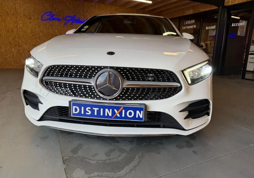 Vue avant d'une Mercedes Classe A 220D AMG Line blanche avec calandre diamantée et phares allumés en intérieur showroom.