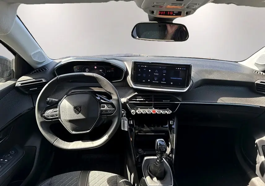 Vue intérieure avant du tableau de bord gris foncé de la Peugeot 208 1.2 100 allure avec volant compact et écran tactile central.