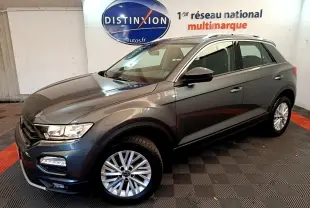Volkswagen T-Roc gris foncé vue 3/4 avant droit, avec jantes alliage et barres de toit argentées visibles.