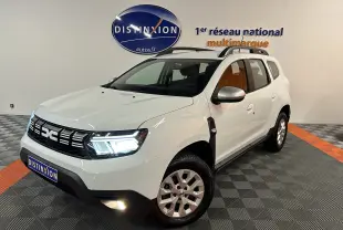 Vue 3/4 avant droite d'un Dacia Duster blanc 2024 avec feux LED allumés et barres de toit noires.