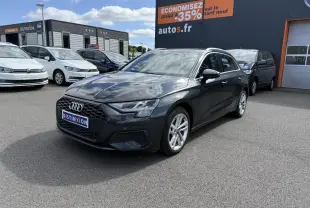 Audi A3 Sportback gris Manhattan métallisé vue 3/4 avant droit sur parking avec enseigne orange en arrière-plan