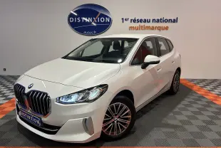 BMW Série 2 Active Tourer blanc vue 3/4 avant droit en intérieur avec calandre imposante et phares allumés.