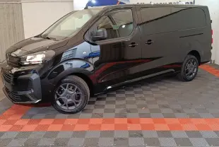 Vue 3/4 avant gauche d’un Citroën Jumpy XL noir avec phares allumés et jantes noires, stationné en intérieur.