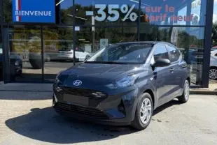 Hyundai i10 gris foncé vue 3/4 avant garée devant une concession Distinxion avec reflet vitré en arrière-plan