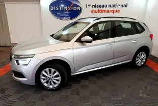 Vue de profil côté gauche d'un Skoda Kamiq gris clair 2022 en showroom avec jantes alliage et barres de toit noires.