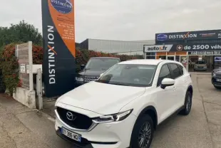 Vue 3/4 avant d'un Mazda CX-5 2.2 SKYACTIV-D 184 blanc, garé devant un concessionnaire Distinxion.