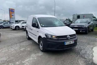Vue 3/4 avant d'un Volkswagen Caddy blanc candy avec pare-chocs noirs, stationné sur un parking sous ciel nuageux.